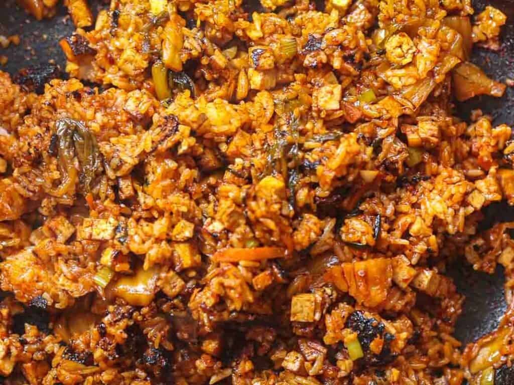 Close-up of Korean vegan kimchi fried rice in a pan. The dish features golden-brown rice mixed with kimchi, diced vegetables, and tofu, all coated in a spicy sauce. The ingredients appear well-cooked with a slightly crispy texture.